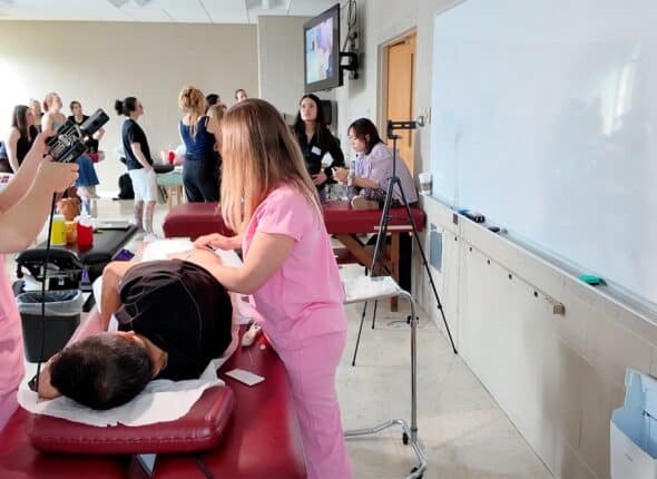 Nursing students in pink scrubs practice patient care on a mannequin in a classroom.