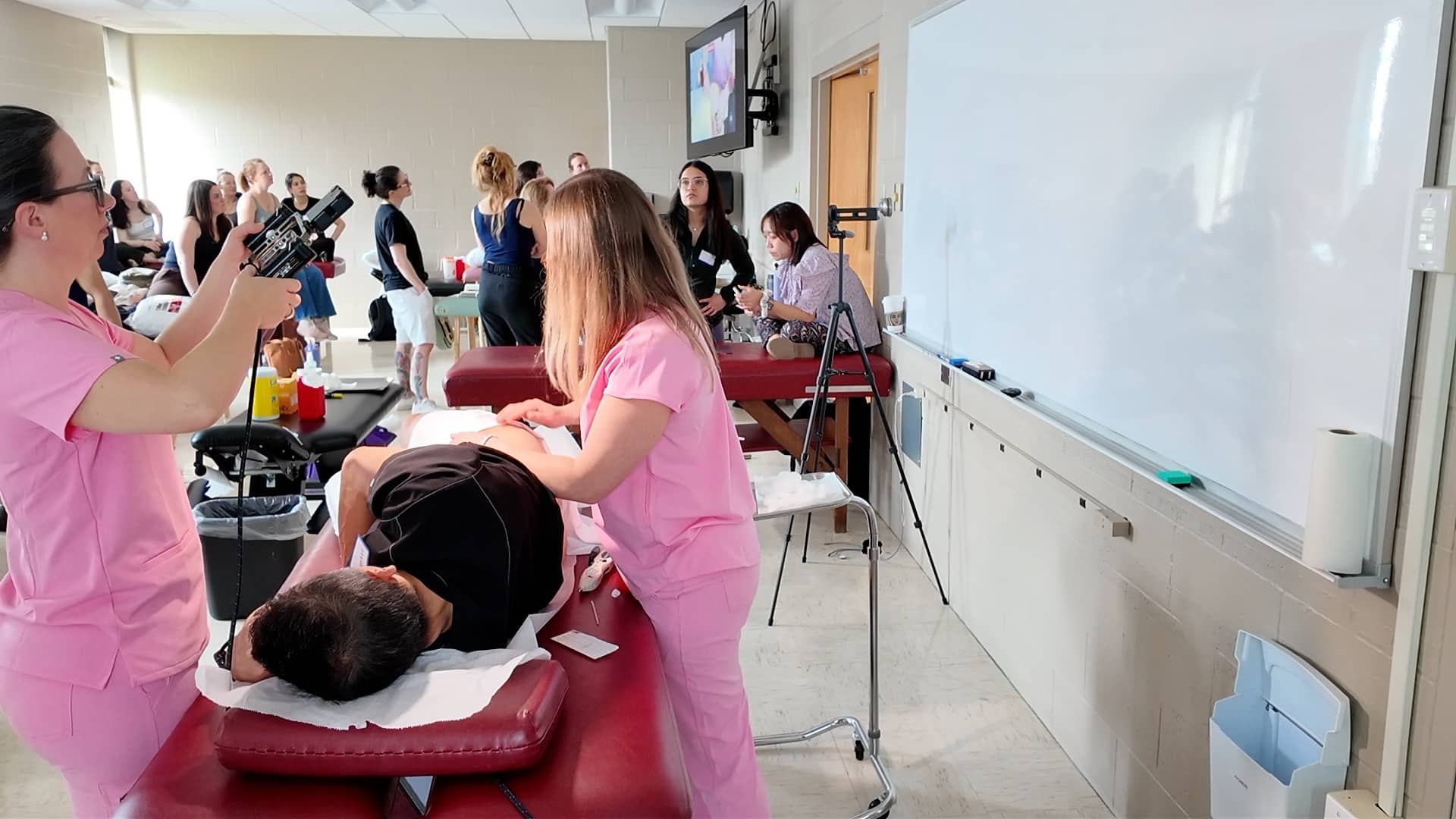 Nursing students in pink scrubs practice patient care on a mannequin in a classroom.