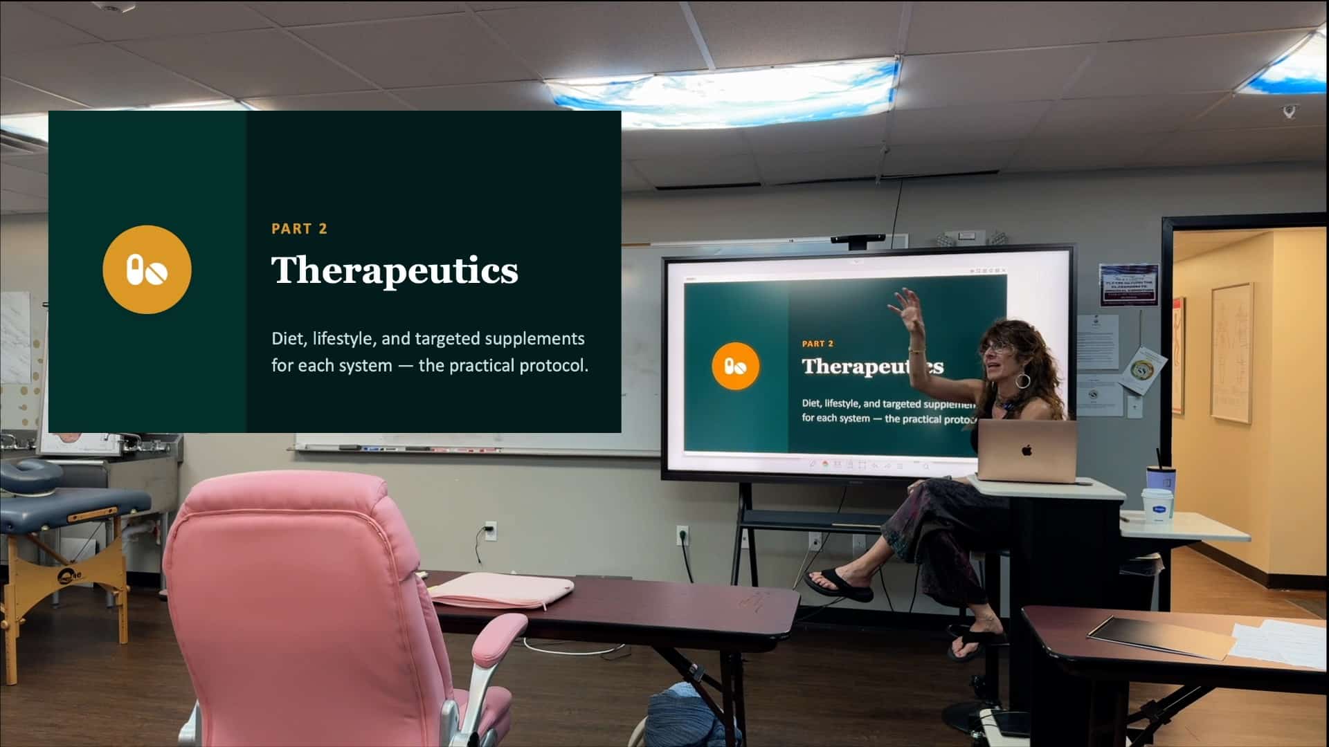 Presentation slide reads 'Therapeutics Part 2' with subtitle about diet, lifestyle, and supplements in a classroom setting. Presenter seated at laptop beside screen.